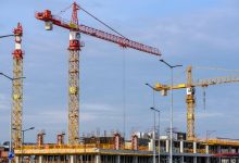 Vários guindastes trabalham em um grande canteiro de obras, sob um céu azul.